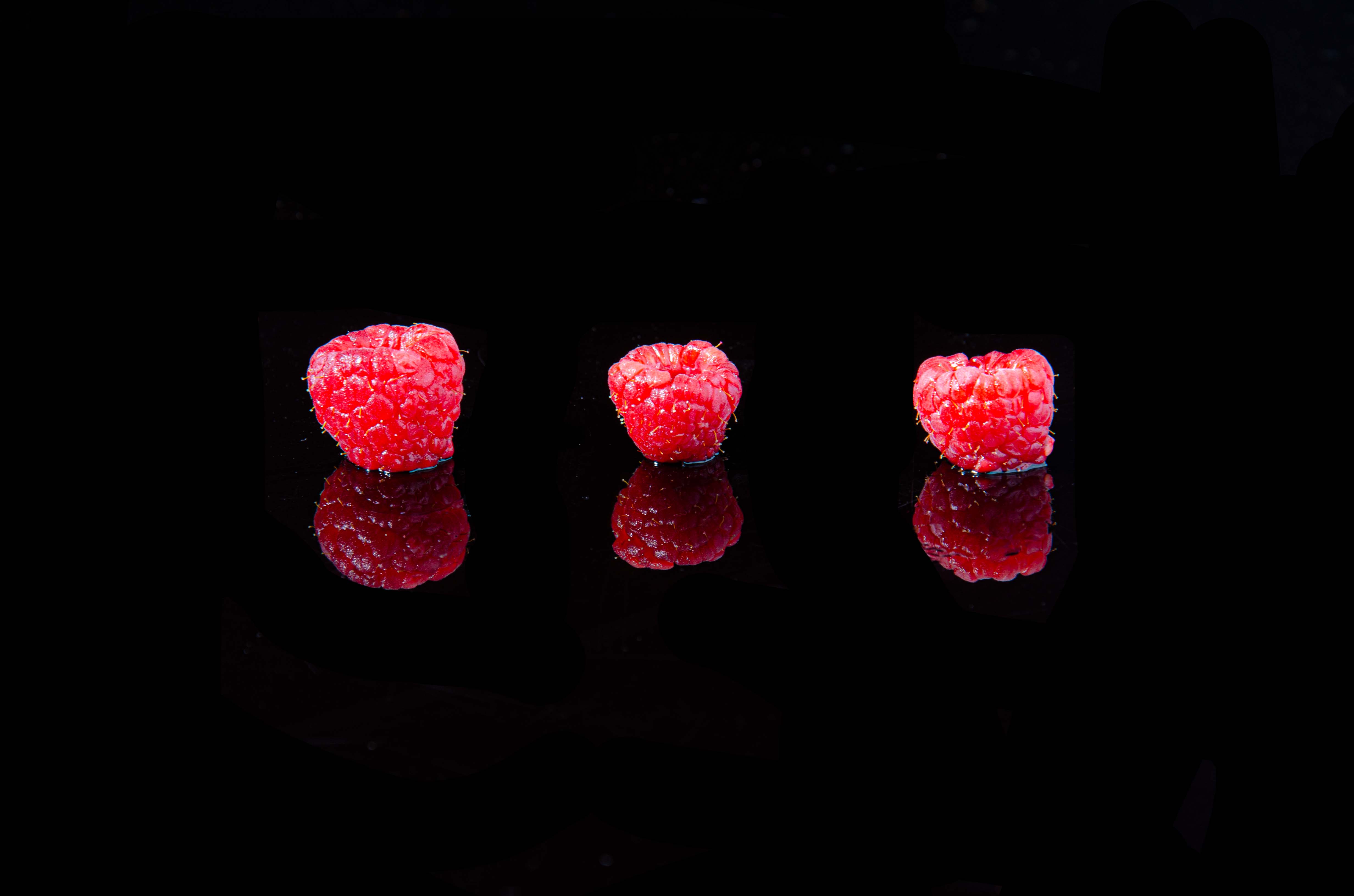 Three rasberries sit in a line on a black reflective table with a black background